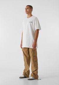 Weißes T-Shirt mit kurzen Ärmeln und schwarzem Text, kombiniert mit khakifarbenen Cargo-Hosen mit sechs Taschen und lockerer Passform sowie grauen Turnschuhen.