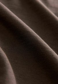 Gros plan sur un tissu marron, présentant une texture lisse, des côtes fines et des variations subtiles de teinte. Le matériau semble doux et flexible.