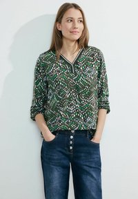 Donkergroene blouse met witte, blauwe en groene stippenpatronen; V-hals ontwerp; lange mouwen; gecombineerd met donkerdenim jeans met knoopdetails.
