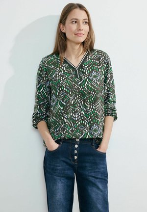 Blouse vert foncé avec des motifs à pois blancs, bleus et verts ; design en V ; manches longues ; associée à un jean en denim foncé avec des détails de boutons.
