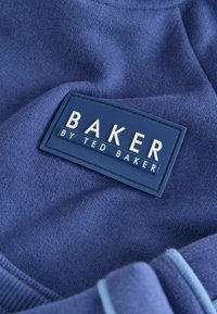 Tumedat sinise kangasriide, millel on ristkülikukujuline silt, millel on valged kirjad "BAKER" ja "by TED BAKER" tekstureeritud taustal.