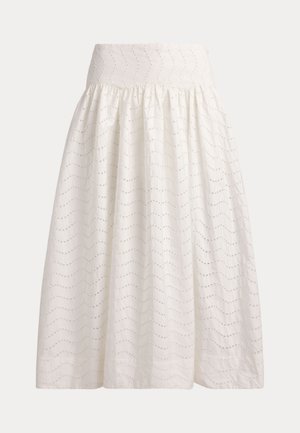 Femme portant un chemisier blanc à manches ballon en œillet et une jupe midi, tenant un sac à main bleu, avec des lunettes de soleil foncées et des sandales orange sur un fond beige.