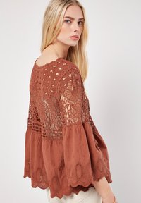 Blouse en crochet marron avec des manches évasées, présentant des motifs en dentelle complexes, un ourlet festonné et une coupe décontractée.