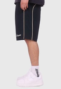 Shorts bleu marine avec passepoil ivoire et logo brodé, associés à des baskets blanches et des chaussettes mi-mollet avec un logo noir.