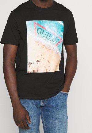 T-shirt en coton noir présentant un grand graphisme triangulaire avec des vagues océanes bleues et une plage de sable, rehaussé par des palmiers. Logo de la marque visible.