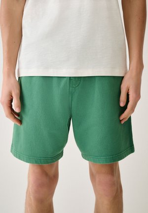 Person trägt grüne Shorts und ein weißes T-Shirt, gezeigt von der Mitte des Oberschenkels bis zur Taille, steht mit entspannten Händen an den Seiten.