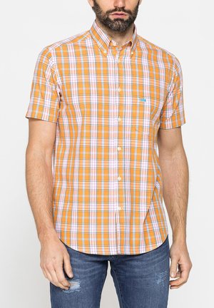 Uomo che indossa una camicia a maniche corte a quadri arancione, bianca e rossa con jeans blu strappati su sfondo uniforme.