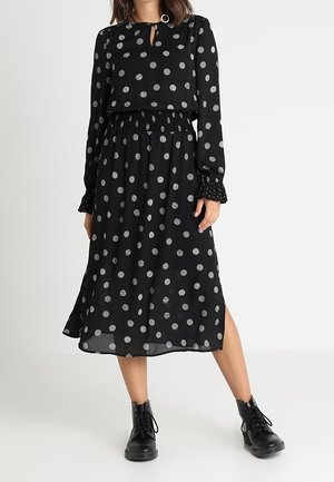 Frau trägt ein schwarzes Midikleid mit weißen Punkten, langen Ärmeln, geraffter Taille, Seitenschlitz und schwarzen Schnür-Stiefeletten.