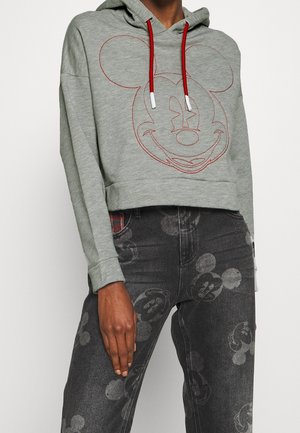 Personne portant un sweat à capuche gris court avec des cordons rouges et un visage brodé de Mickey Mouse, associé à un jean foncé avec des impressions délavées de Mickey Mouse.