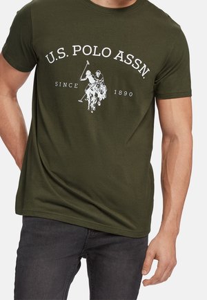 Mand iført olivengrøn T-shirt med hvid U.S. Polo Assn.-tekst og logo med to polospillere på heste, der holder køller.