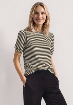 Femme souriante aux cheveux blonds mi-longs, portant un haut à manches courtes rayé noir et blanc et un pantalon noir, les mains dans les poches.