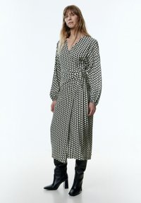 Langes Kleid mit einem schwarz-weißen geometrischen Muster, das ein Wickeldesign und ballonförmige Ärmel aufweist, kombiniert mit knielangen schwarzen Stiefeln mit Absätzen.