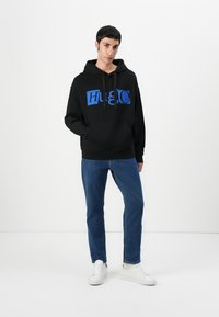 Hanorac negru din material moale, cu un mare logo albastru "Hug" pe partea din față, cu o buzunar tip cangur și glugă cu șnur. Asortat cu blugi albaștri și adidași albi.