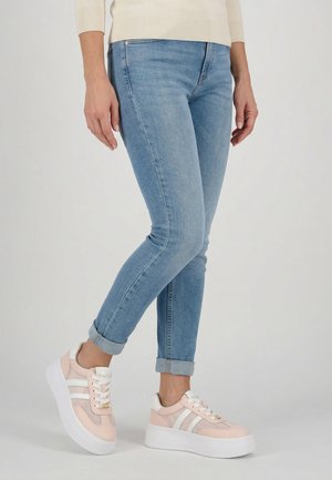 Osoba nosząca jasnoniebieskie wąskie dżinsy z podwiniętymi nogawkami oraz różowo-białe sneakersy na platformie, stojąca z jedną lekko uniesioną stopą.