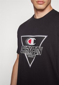 Musta t-paita, joka on valmistettu puuvillasta, ja jossa on suuri valkoinen ja punainen "Champion" -logo kolmiossa, tekstillä "ATHLETICWEAR 1992" alla.