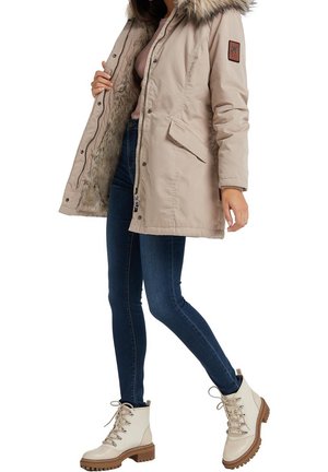 Femme portant un parka beige avec capuche en fourrure, un jean skinny foncé et des bottes claires à lacets, posant avec une jambe légèrement levée.
