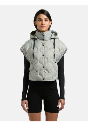 Kvinde iført kort, quiltet grå hættevest med sorte snore over sort langærmet top og shorts, stående mod en ensfarvet baggrund.