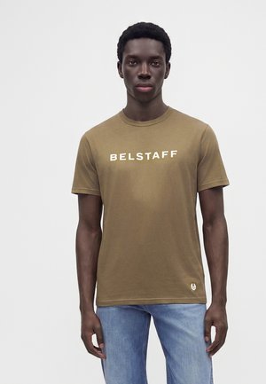 Młody mężczyzna o ciemnej karnacji, ubrany w oliwkową koszulkę Belstaff i jasnoniebieskie dżinsy, stoi na tle jednolitego jasnego tła.