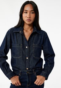 Mørkeblå denimskjorte med spids krave, brystlommer og knaplukning foran. Har gyldne syninger og en afslappet pasform.