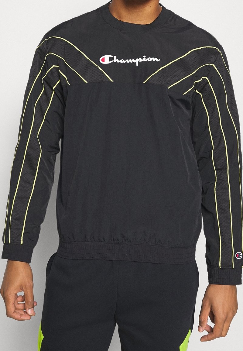 Svart atletisk jacka med gula randdetaljer, tillverkad av lättviktig material. Har rund hals och elastisk midja; logotyp på bröstet.