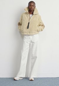 Veste en fausse fourrure beige avec un col haute et une fermeture éclair, associée à un pantalon blanc à jambe évasée et des chaussures de couleur claire.