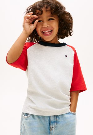 Enfant souriant aux cheveux bouclés portant un T-shirt blanc à manches rouges et col bleu, assorti à un short en jean bleu clair.