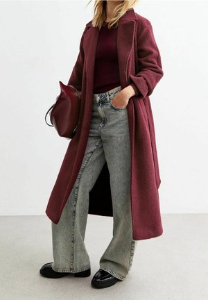Femme portant un long manteau bordeaux, un pull assorti, un jean gris à jambes larges, des chaussures noires et tenant un sac à main bordeaux.