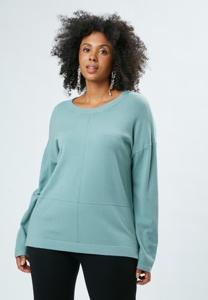 Suéter verde azulado oversize con cuello redondo, mangas largas y tela texturizada con detalles de panel. Combinado con leggings negros.