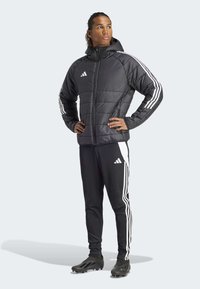 Svart polstret jakke med hette, med hvite striper på ermene; sammen med svarte bukser for treningsdrakt med hvite detaljer og svarte joggesko.