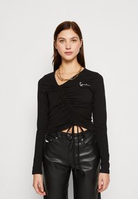 Sort langærmet top og højtaljede læderbukser. Toppen har rynk og snøredetaljer, med en hvid logoaccent på brystet.