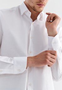 Biała koszula zapinana na guziki, z szerokim kołnierzem, długimi rękawami i okrągłymi mankietami. Tkanina jest gładka, o jednolitej strukturze i subtelnym połysku.