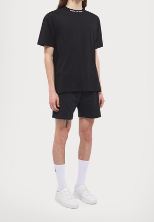 Sort bomulds T-shirt med rund hals, der har teksten "VISION OF SUPER", parret med sorte shorts, hvide sokker og hvide sneakers.