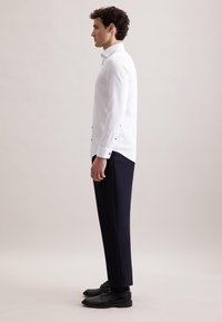 Witte blouse met lange mouwen, voorzien van zwarte hardware accenten, gecombineerd met donkerblauwe broek en zwarte nette schoenen. Zijaanzicht.