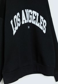 Sweat-shirt noir avec "LOS ANGELES" imprimé en grandes lettres blanches incurvées sur la poitrine.