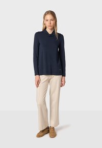 Marineblaues Langarmoberteil mit einem Schalkragen, kombiniert mit beigen Schlaghosen und braunen Wildledersneakern, schafft eine kontrastierende neutrale Farbpalette.