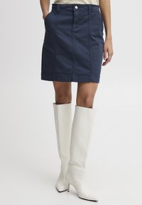 Jupe en coton bleu marine avec des coutures à l'avant et des poches, associée à des bottes blanches ajustées montant jusqu'à la cuisse, présentant une surface texturée et des pointes relevées.