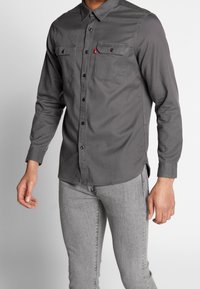 Grå skjorta med två bröstfickor, svarta knappar, långa ärmar och en något böjd nederkant, matchad med ljusgrå jeans.