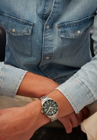 Personne portant une chemise en denim bleu clair et une montre bracelet argentée avec un cadran vert foncé au poignet gauche, les mains reposant dans les genoux.