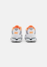 Un par de zapatillas Nike blancas y grises con acentos naranjas, vistas desde la parte de atrás sobre un fondo gris claro liso.