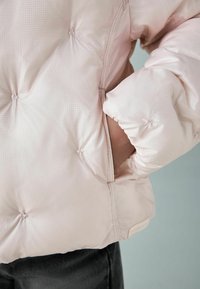 Lichtroze pufferjack met een gestructureerd oppervlak, voorzien van een zijzak en verhardde mouwuiteinden, vastgezet met decoratieve stiksels.