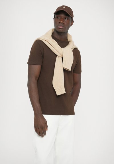 Barna pamut póló rövid ujjúval, hozzá illő bézs kötött pulóver vállra vetve és fehér nadrág. Barna sapka logóval.