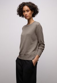 Pull en maille marron clair avec un col rond, des poignets et un ourlet côtelés, associé à un pantalon noir ajusté. Texture lisse, design minimaliste.