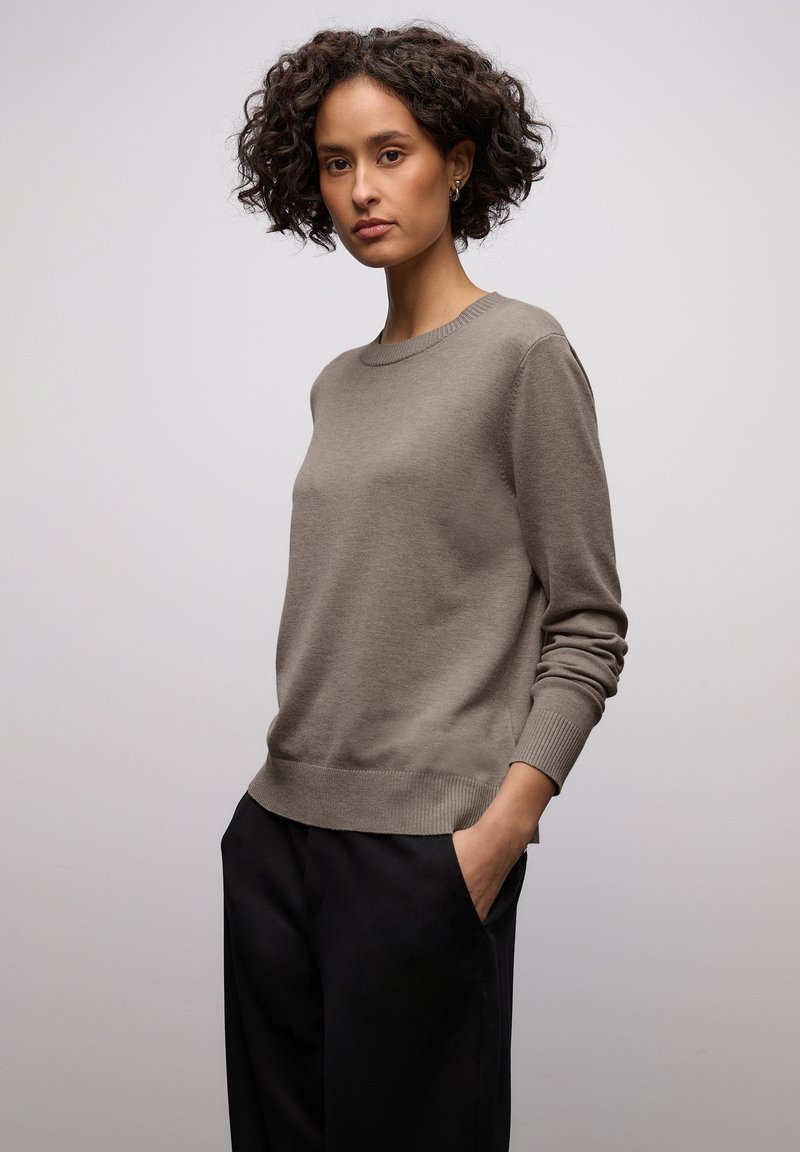 Pull en maille marron clair avec un col rond, des poignets et un ourlet côtelés, associé à un pantalon noir ajusté. Texture lisse, design minimaliste.