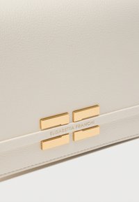 Λευκή δερμάτινη τσάντα χειρός με ανάγλυφο λογότυπο, χρυσές λεπτομέρειες και μινιμαλιστικό σχέδιο που περιλαμβάνει οριζόντιες και κάθετες ορθογώνιες λεπτομέρειες.
