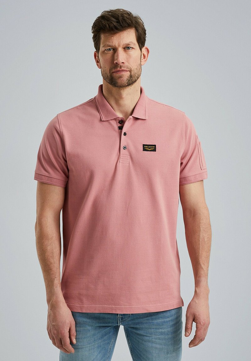 Light pink polo shirt with short sleeves, three-button placket, and a small black label on the left chest. Worn with blue jeans.