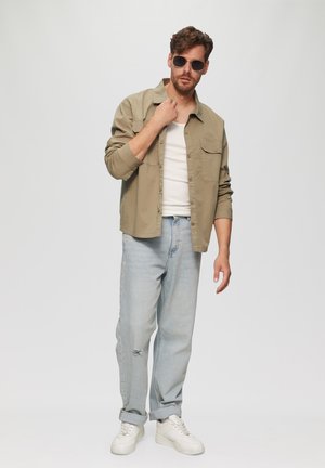 Lightweight beige button-up shirt over a white tank, paired with light blue jeans and white sneakers. Accessories include dark sunglasses.
