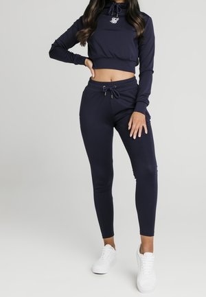 Kobieta ubrana w granatową krótką bluzę z kapturem i dopasowane dresy oraz białe tenisówki, pozująca na tle jednolitego jasnoszarego tła.