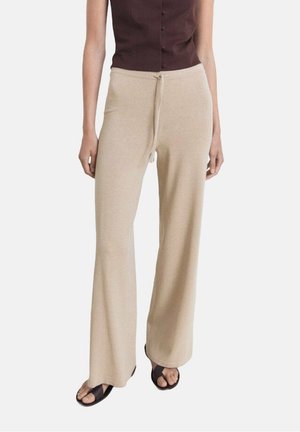 Kobieta ubrana w beżowe spodnie o szerokich nogawkach z wiązanym paskiem w talii, w połączeniu z ciemnobrązową, bezrękawnikową bluzką zapinaną na guziki oraz czarnymi sandałami.