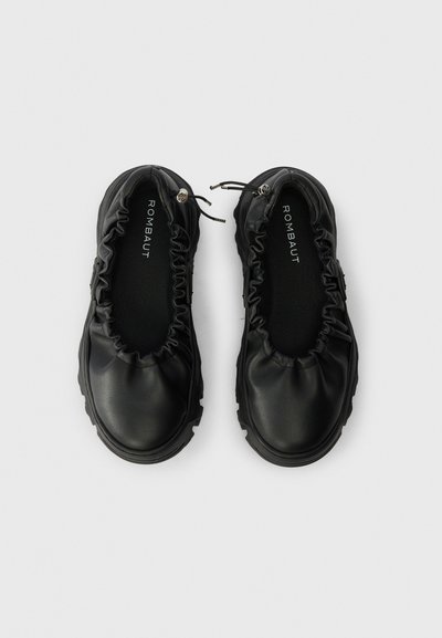 Un par de zapatos negros slip-on con ribete elástico fruncido y suelas con taco, vistos desde arriba sobre un fondo plano.