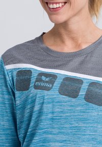 Hellblaues Sport-T-Shirt mit grauen Akzenten, strukturiertem Ausschnitt und horizontalem Grafikdesign mit schwarzen Streifen und Logo.